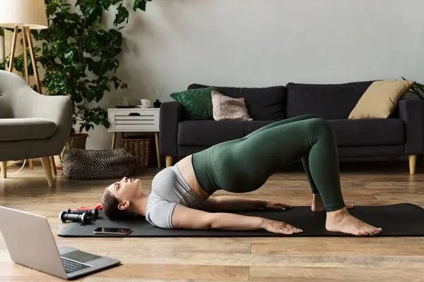 Una mujer deportiva se ejercita en casa, centrándose en su forma mientras realiza una pose de puente en una esterilla de yoga. - foto de stock