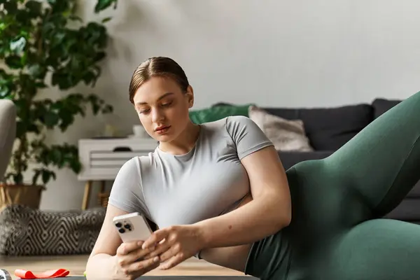 Una mujer joven se ejercita en casa en ropa activa elegante mientras se centra en su teléfono inteligente. - foto de stock