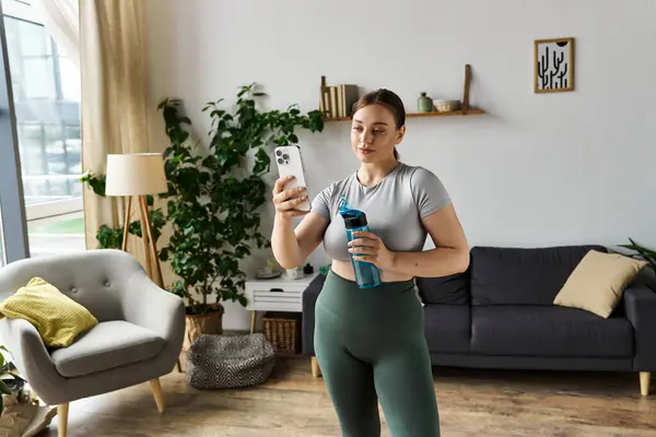 Una donna sportiva in un crop top e leggings gode di un allenamento a casa, prendendo una pausa per controllare il suo telefono. — Foto stock