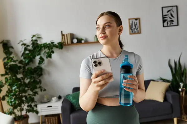 Eine junge Frau übt zu Hause, hält eine Wasserflasche und ein Telefon in der Hand, um ihre Fitness zu verbessern. — Stockfoto