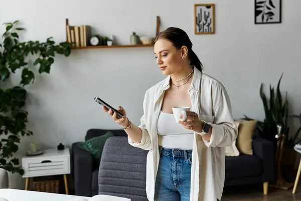 Una giovane donna bilancia casualmente il suo tempo tra il telefono e il caffè mentre lavora da casa. — Foto stock