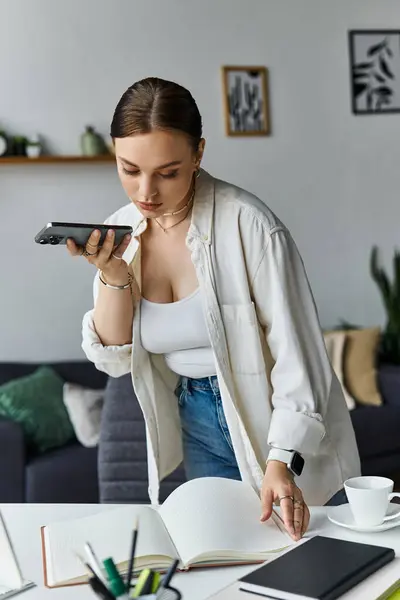 Eine junge Frau in lässiger Kleidung spricht in ihr Telefon, während sie Notizen am heimischen Arbeitsplatz durchsieht. — Stockfoto
