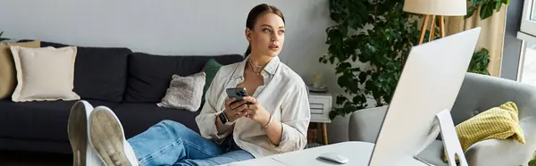 Молода жінка комфортно працює з дому, сидячи розслабленим зі своїм телефоном, взаємодіючи з її комп'ютером. — стокове фото