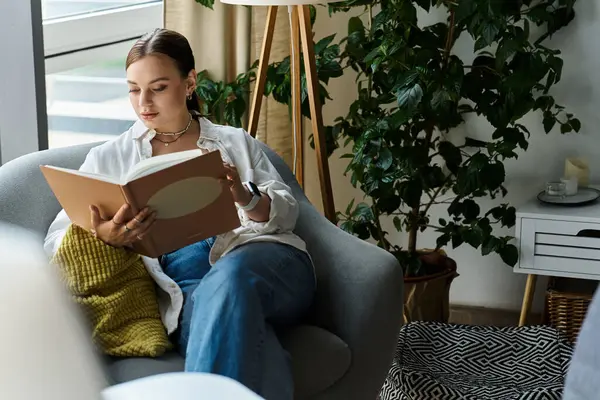 Una joven se relaja en su sofá, absorta en un libro mientras disfruta de las comodidades del hogar. - foto de stock