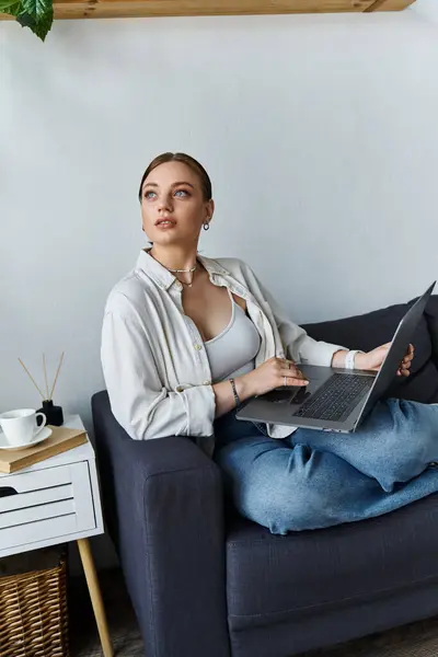 A young woman casually dressed sits comfortably on her couch, working intently on her laptop. — Stock Photo