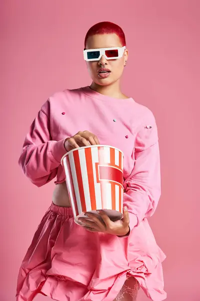 Die junge Frau präsentiert ihre einzigartige Mode, hält einen gestreiften Eimer mit Popcorn vor einem verspielten rosa Hintergrund. — Stockfoto