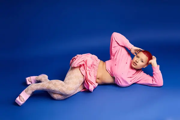 A confident young woman with pink hair and a nose piercing poses stylishly against a blue backdrop. — Stock Photo