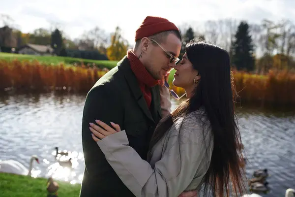 Two people share a tender moment by a serene lake, adorned in cozy fall outfits. — Stock Photo
