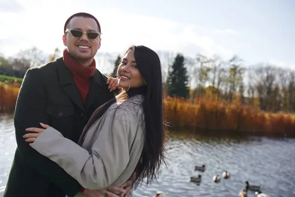 A couple in warm, cozy attire embraces lovingly by a serene lake, surrounded by autumn beauty. — Stock Photo