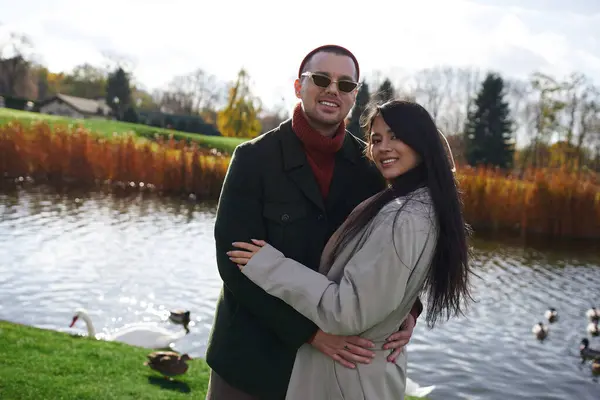 A couple in stylish autumn attire joyfully poses together by a peaceful lake, enjoying the moment. — Stock Photo