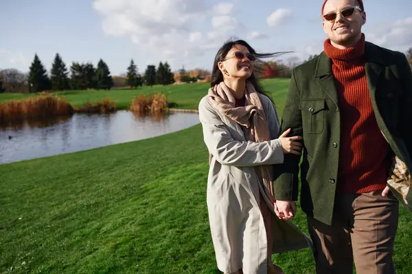 A couple strolls hand in hand by a shimmering pond, wrapped in warm autumn clothing, smiling. — Stock Photo