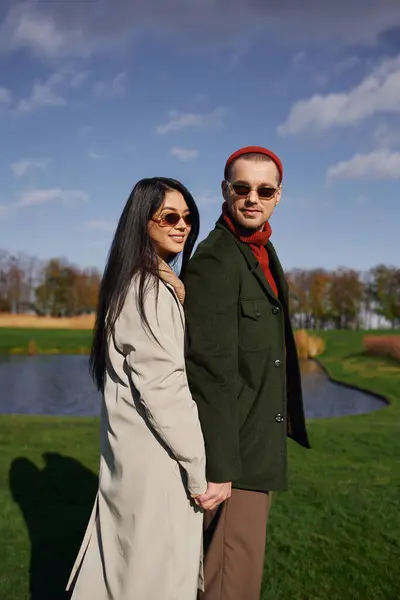A couple enjoys a tender moment, hand in hand, dressed in warm autumn attire near a peaceful pond. — Stock Photo