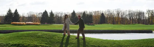 Una pareja cariñosa camina de la mano a lo largo de un exuberante lago verde, envuelto en acogedoras capas de otoño. — Stock Photo