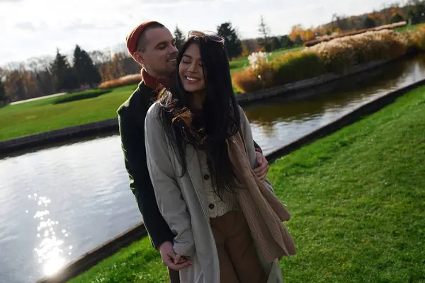 A couple embraces warmly in autumn attire, sharing smiles by serene waters under golden trees. — Stock Photo