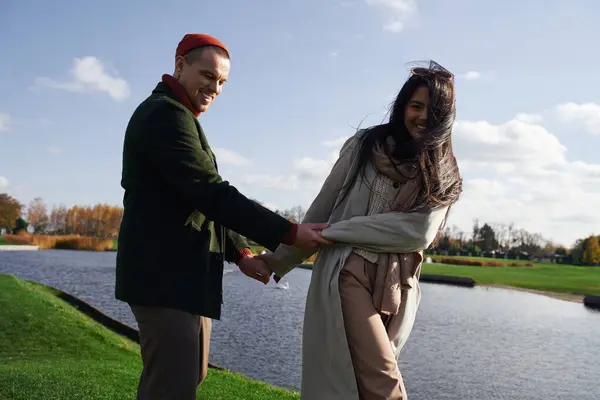 Two people share laughter and joy while holding hands near a peaceful lake on an autumn day. — Stock Photo