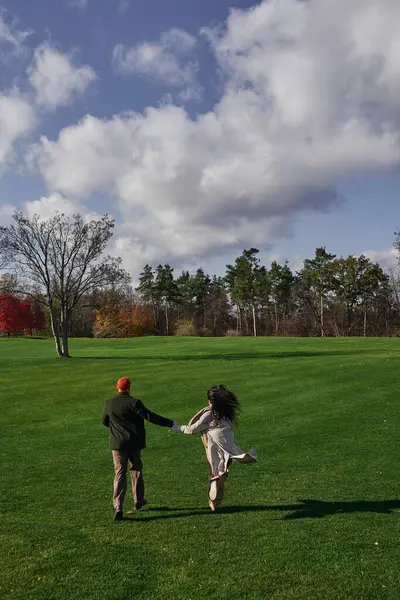 A loving couple embraces the autumn chill, laughing as they sprint across a lush green field. — Stock Photo