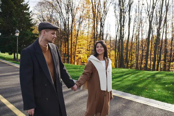 A couple dressed in cozy attire walks hand in hand, sharing smiles amidst autumn foliage. — Foto stock