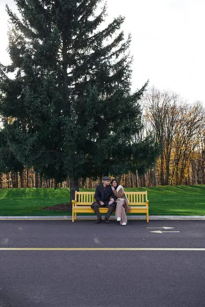 A couple enjoys a warm moment on a yellow bench surrounded by autumn trees and greenery. — Stock Photo