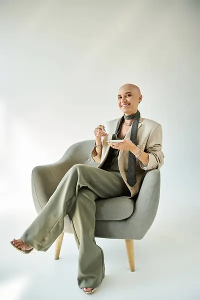 A young bald woman with alopecia sits elegantly in a cozy chair, savoring her tea. — Stock Photo