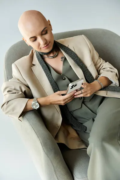 A confident young woman with alopecia relaxes in a stylish chair, engaged with her smartphone. — Stock Photo