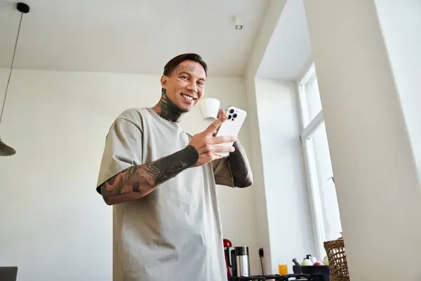 A cheerful young man in a casual t shirt happily interacts with his phone while holding a cup. — Stock Photo