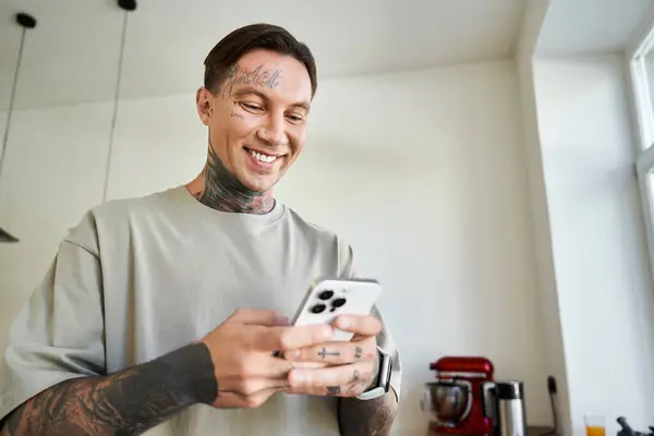 Um homem jovem e atraente relaxa em casa, sorrindo enquanto navega em seu smartphone. — Fotografia de Stock