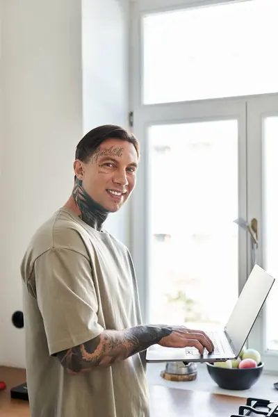 A young man with tattoos smiles while using his laptop in a modern home setting filled with light. — Stock Photo