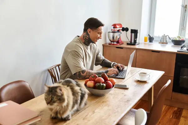 The young man engages in focused work on his laptop, surrounded by fresh fruit and a curious cat. — Stock Photo