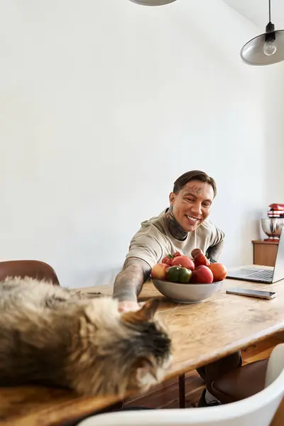 A cheerful young man reaches out to his cat while surrounded by a bowl of fresh fruit at home. — Stock Photo