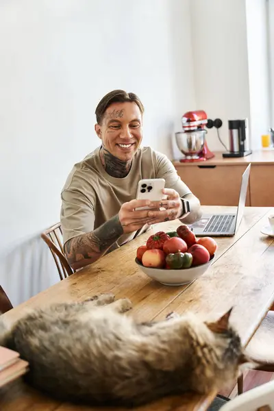 A young man smiles at his smartphone at home, with a cat and fruit on the table. — Stock Photo