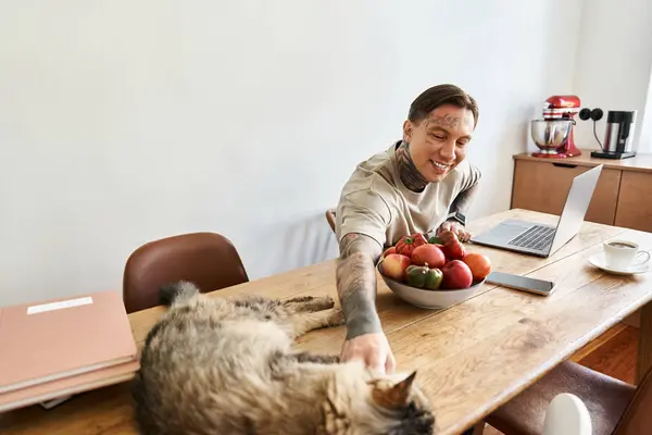 Smiling man interacts with his cat while working at a wooden table filled with fresh fruit. — Stock Photo