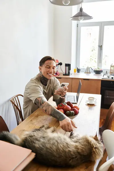 A cheerful young man interacts with his cat at a cluttered kitchen table surrounded by fruits. — Stock Photo