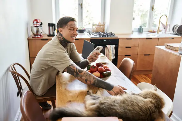 Un joven sonríe mientras se sienta en la mesa de la cocina, disfrutando del tiempo con su gato y teléfono. - foto de stock