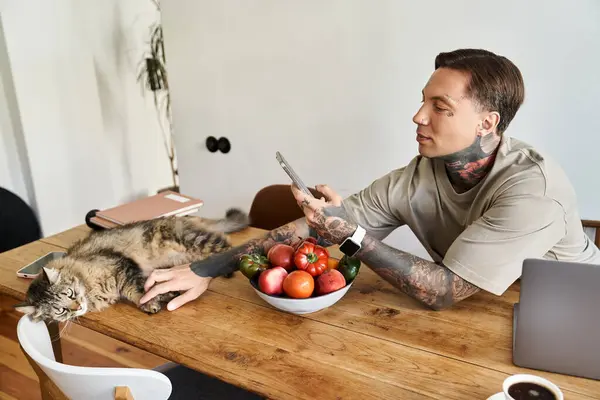 Ein junger Mann betätigt sich mit seinem Handy, während er zu Hause neben seiner entspannten Katze und frischen Produkten faulenzt. — Stockfoto