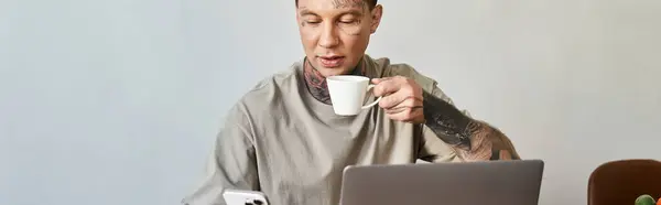 A young man enjoys coffee at home, absorbed in his phone and laptop, creating a cozy vibe. — Stock Photo
