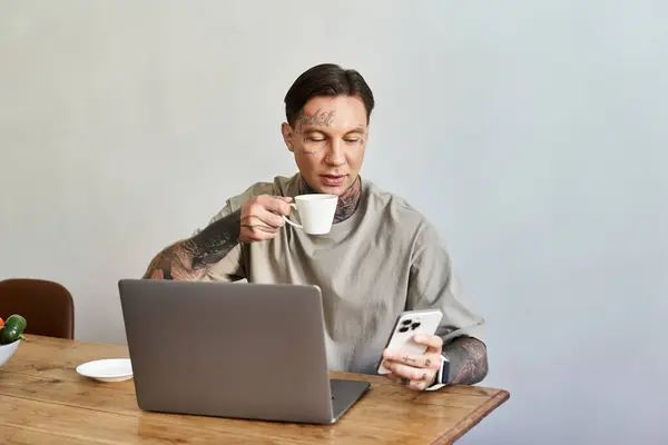 A young handsome man sips coffee, checking his phone while sitting at a wooden table. — Stock Photo