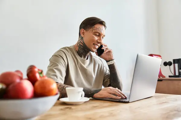 The young man sits at a wooden table surrounded by fresh fruits while engaged in a phone call. — Stock Photo