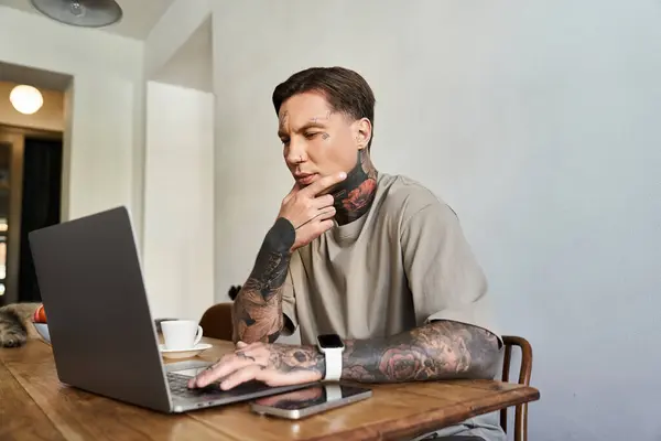 A focused young man with tattoos engages with his laptop at home while enjoying coffee. — Stock Photo