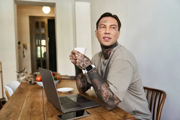 A stylish young man with tattoos savors coffee while engaging with his laptop in a warm home. — Stock Photo