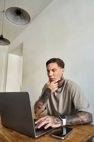 Un hombre joven y tatuado se involucra pensativamente con su portátil en casa, inmerso en el trabajo. - foto de stock