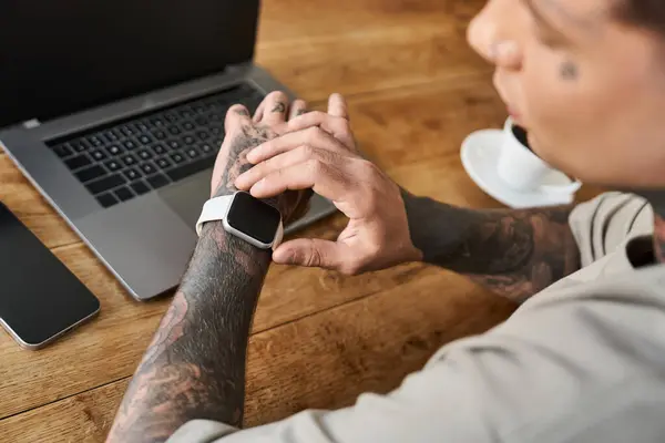 Ein stilvoller junger Mann beobachtet aufmerksam seine Smartwatch, während er am Schreibtisch sitzt. — Stockfoto