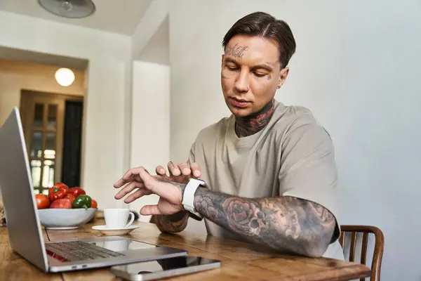 A young, tattooed man intently focuses on his laptop while glancing at his smartwatch. — Stock Photo