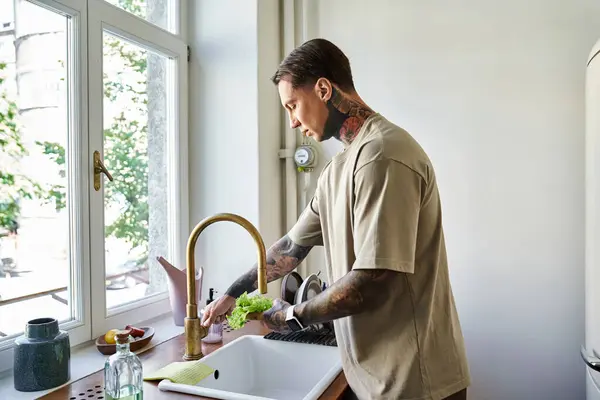 A young man attentively washes fresh vegetables at his cozy kitchen sink, surrounded by sunlight. — Stock Photo
