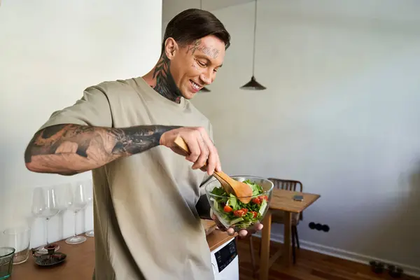 A young handsome man smiles as he mixes a vibrant salad in his cozy kitchen at home. — Stock Photo