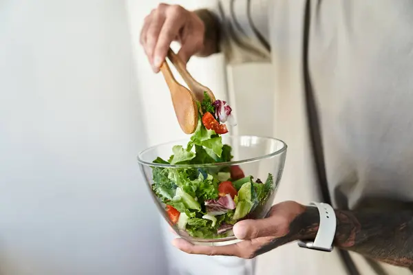 This young man skillfully tosses a vibrant salad in a glass bowl, blending fresh ingredients. — Stock Photo