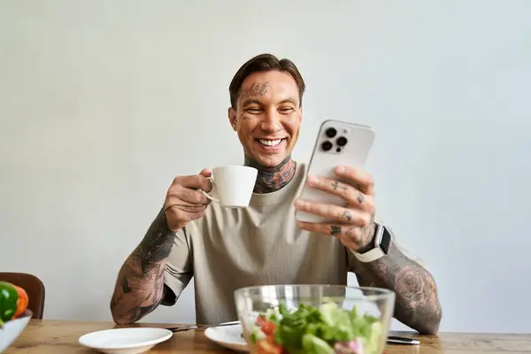 A cheerful young man is sipping coffee and smiling at his smartphone during breakfast at home. — Stock Photo
