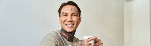 Dans une pièce calme, un jeune homme sourit avec une boisson chaude, affichant fièrement ses tatouages. — Photo de stock