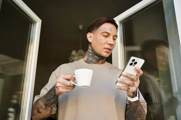 A handsome young man stands on his balcony, sipping coffee and browsing his phone, relaxed at home. — Stock Photo