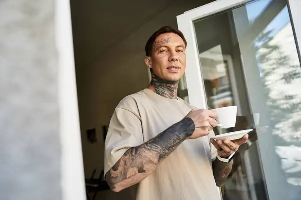 A young man with tattoos relaxes at home, sipping coffee near an open window. — Stock Photo