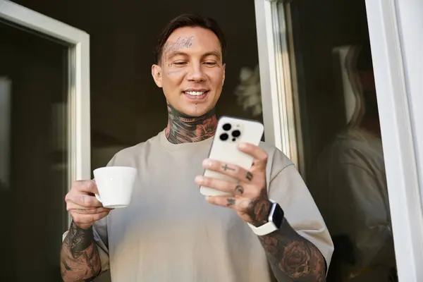 A young man with tattoos smiles as he sips coffee and looks at his phone on a cozy morning. — Stock Photo